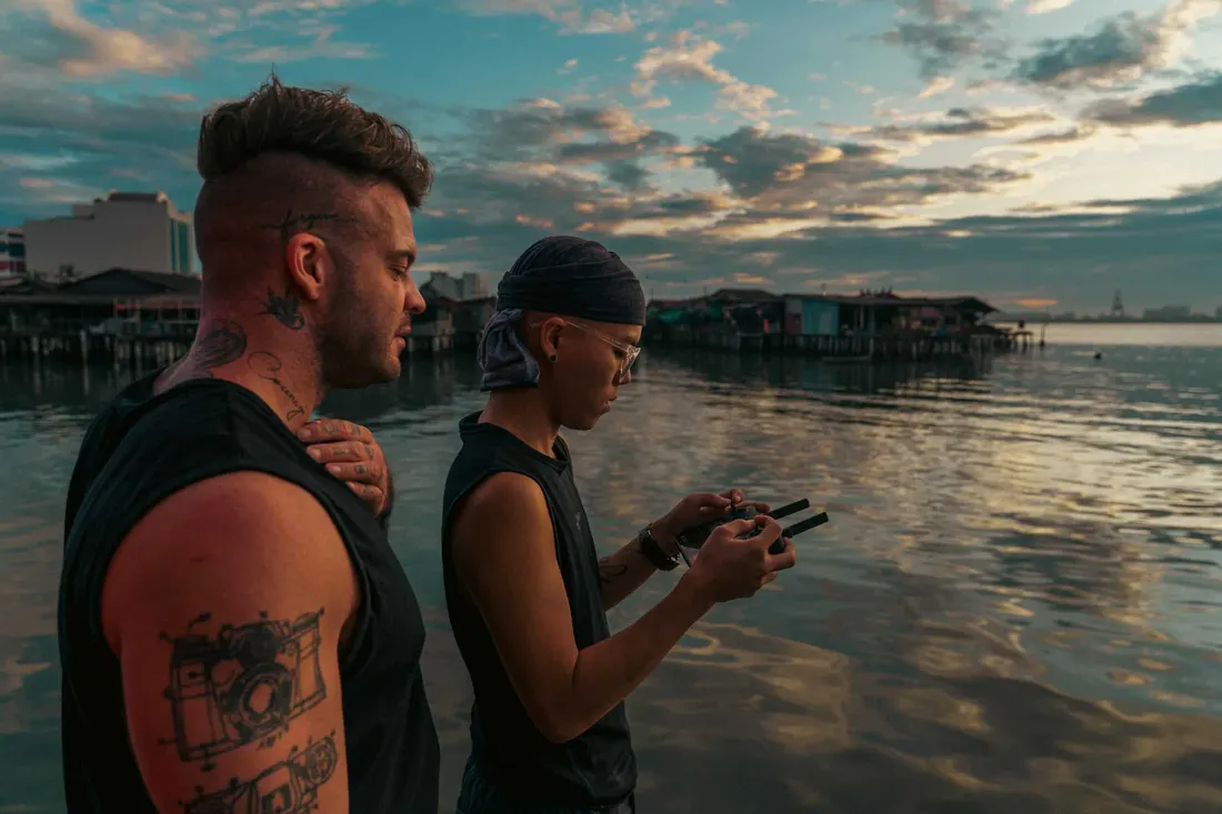 Two crew members at dusk beside a stilt-house waterfront, one flying a drone from a handheld controller.