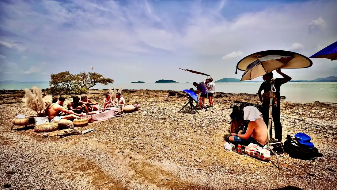 A wide shot of a production crew filming cast members seated on a rocky coastal shoreline, with camera and lighting kit set up nearby.