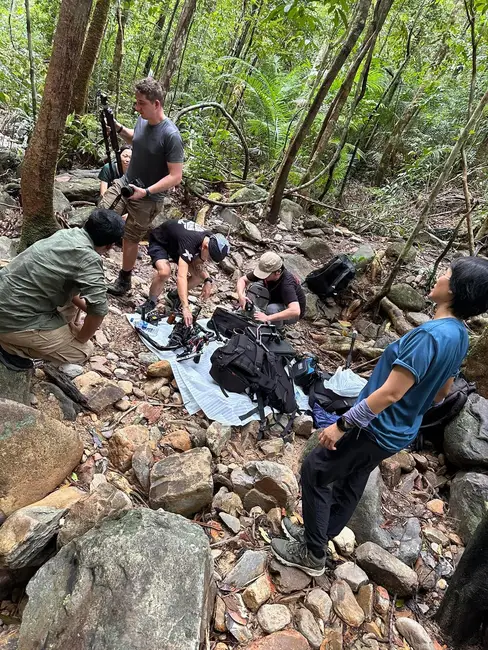 A crew gathers on a rocky jungle trail to unpack and assemble camera equipment laid out on a groundsheet.