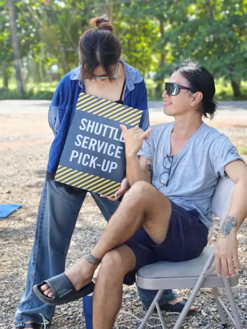 Two crew members at a roadside holding area with a yellow and black Shuttle Service Pick-Up sign, one seated on a folding chair.