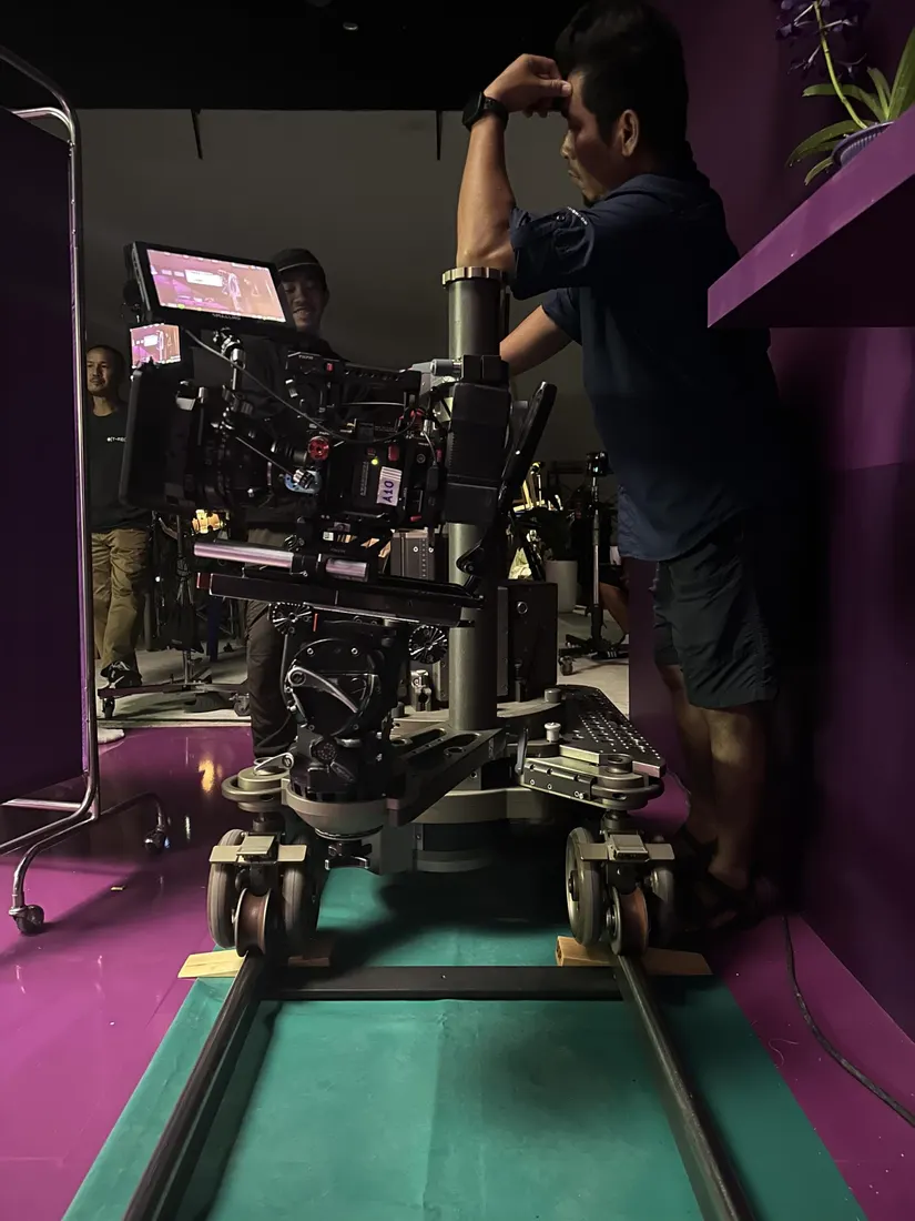 A camera rig on a dolly track inside a magenta-lit set, with a crew member checking the shot.
