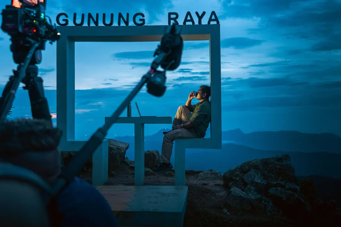 A crew filming at a mountain viewpoint at dusk, camera and boom rigged around a white frame structure lettered Gunung Raya, with the ridgeline behind.