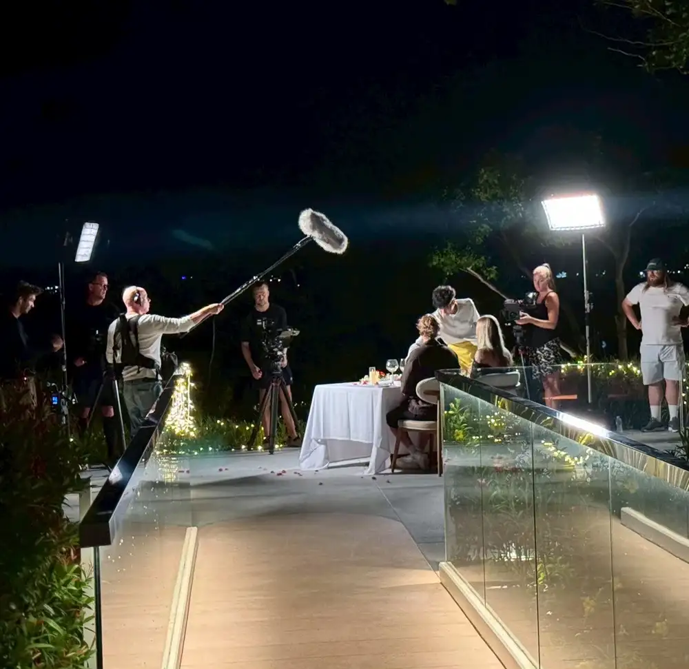 A night shoot on a lit terrace, a boom operator and camera crew working around a dressed dinner table.