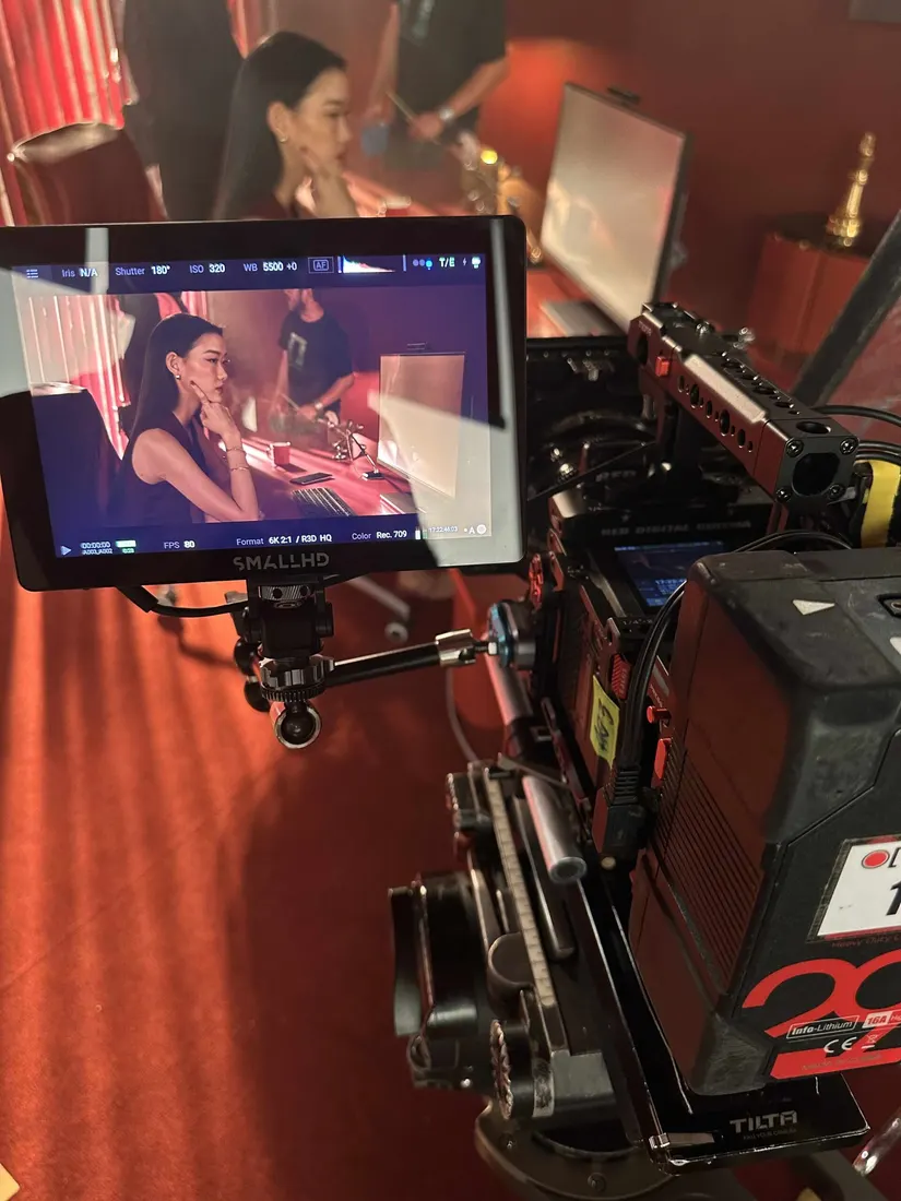 A cinema camera monitor shows a seated talent lit in red on set, with the operator's rig in the foreground.