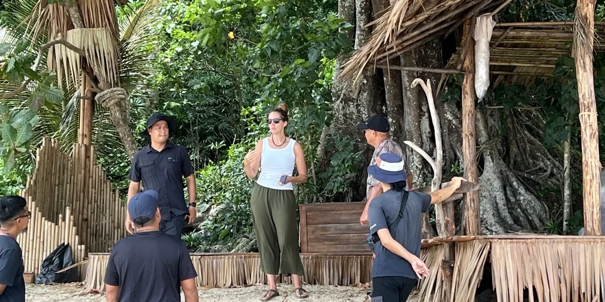 Crew on a beach location beside a bamboo and thatch set built into the treeline, talking through the shot.