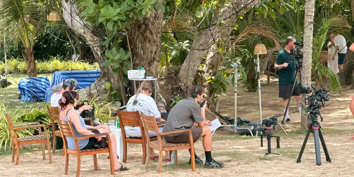 Crew sitting in the shade of large trees between takes, with cameras on tripods standing by on the sand.